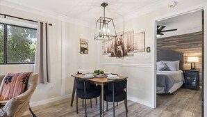 Una habitación con una mesa de comedor, sillas, una cama, un ventilador de techo y una ventana con cortinas.