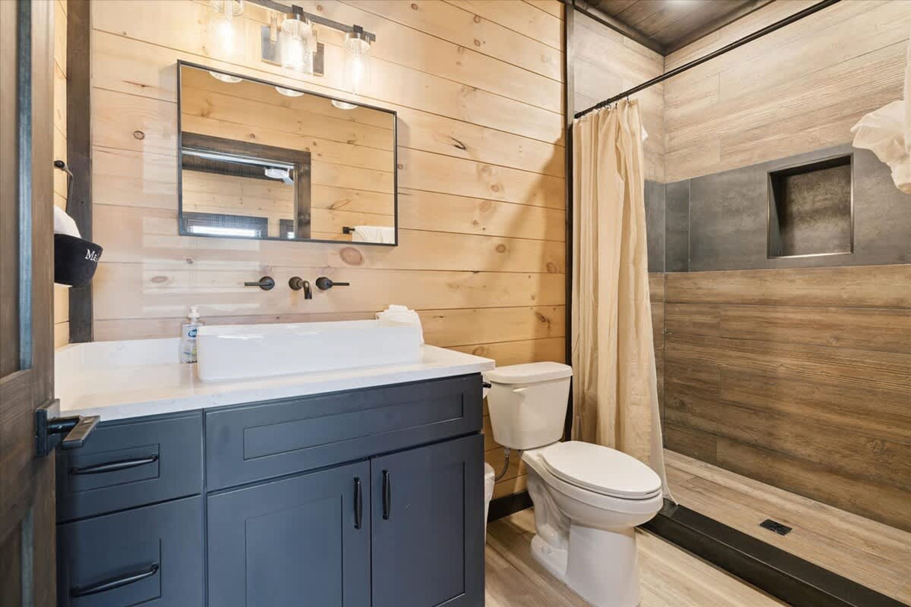 Une salle de bain aux murs lambrissés, avec un lavabo blanc, des meubles sombres, des toilettes et un rideau de douche.