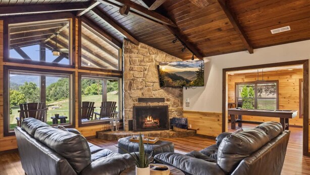 Ein gemütliches Wohnzimmer mit Ledersofas, einem Kamin aus Stein und großen Fenstern mit Blick ins Freie.