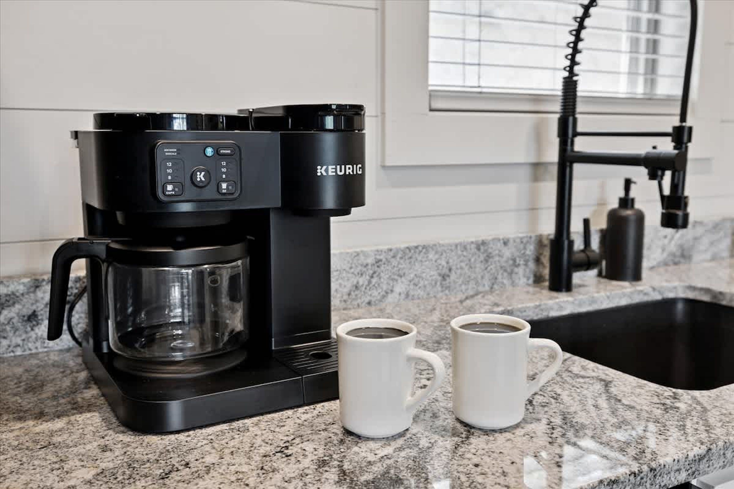 Een Keurig koffiezetapparaat op een granieten aanrecht met twee witte mokken ernaast.