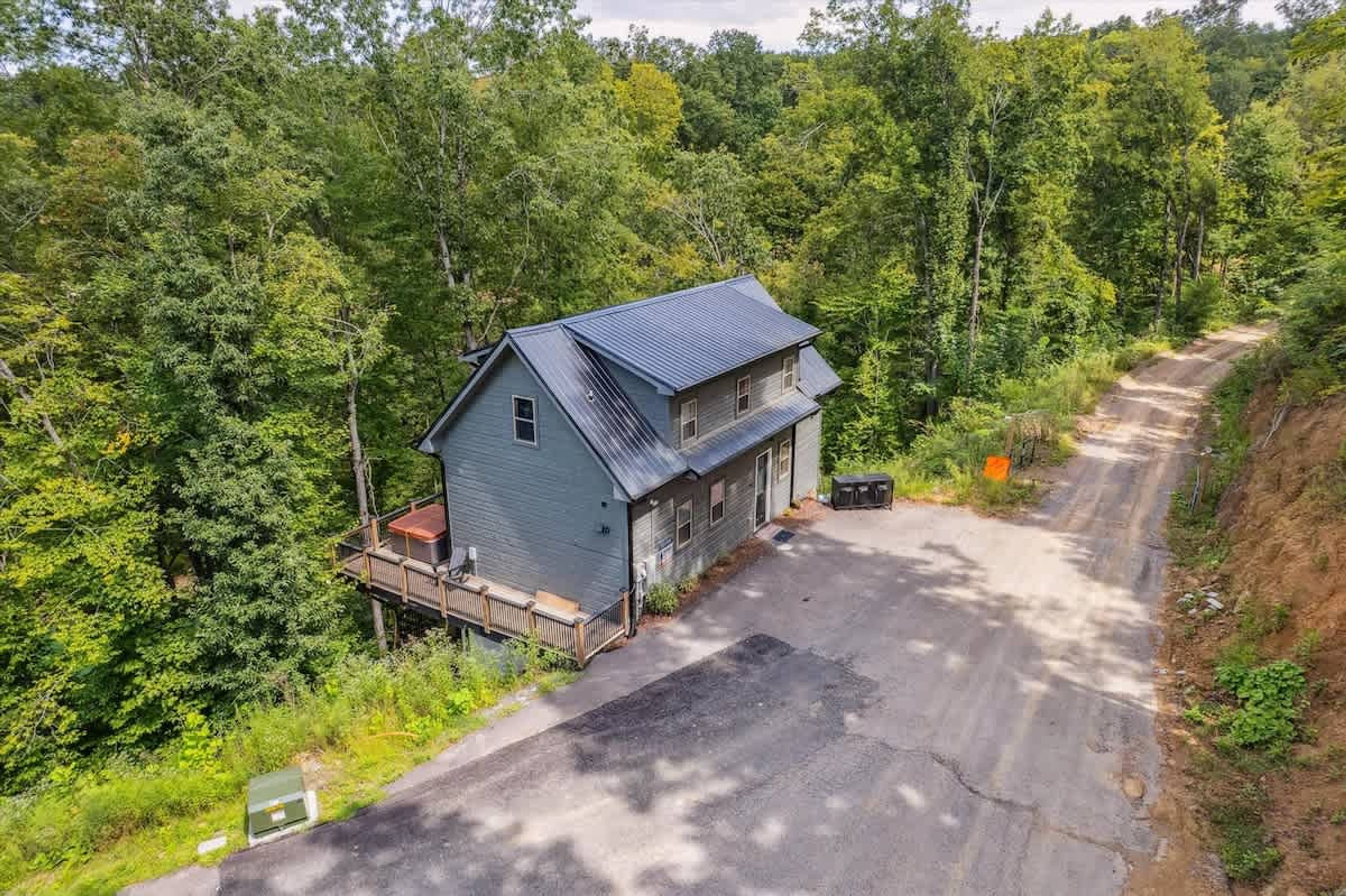 Une maison à deux étages, avec un toit en métal, entourée d’une forêt dense, et un chemin de terre qui y mène.