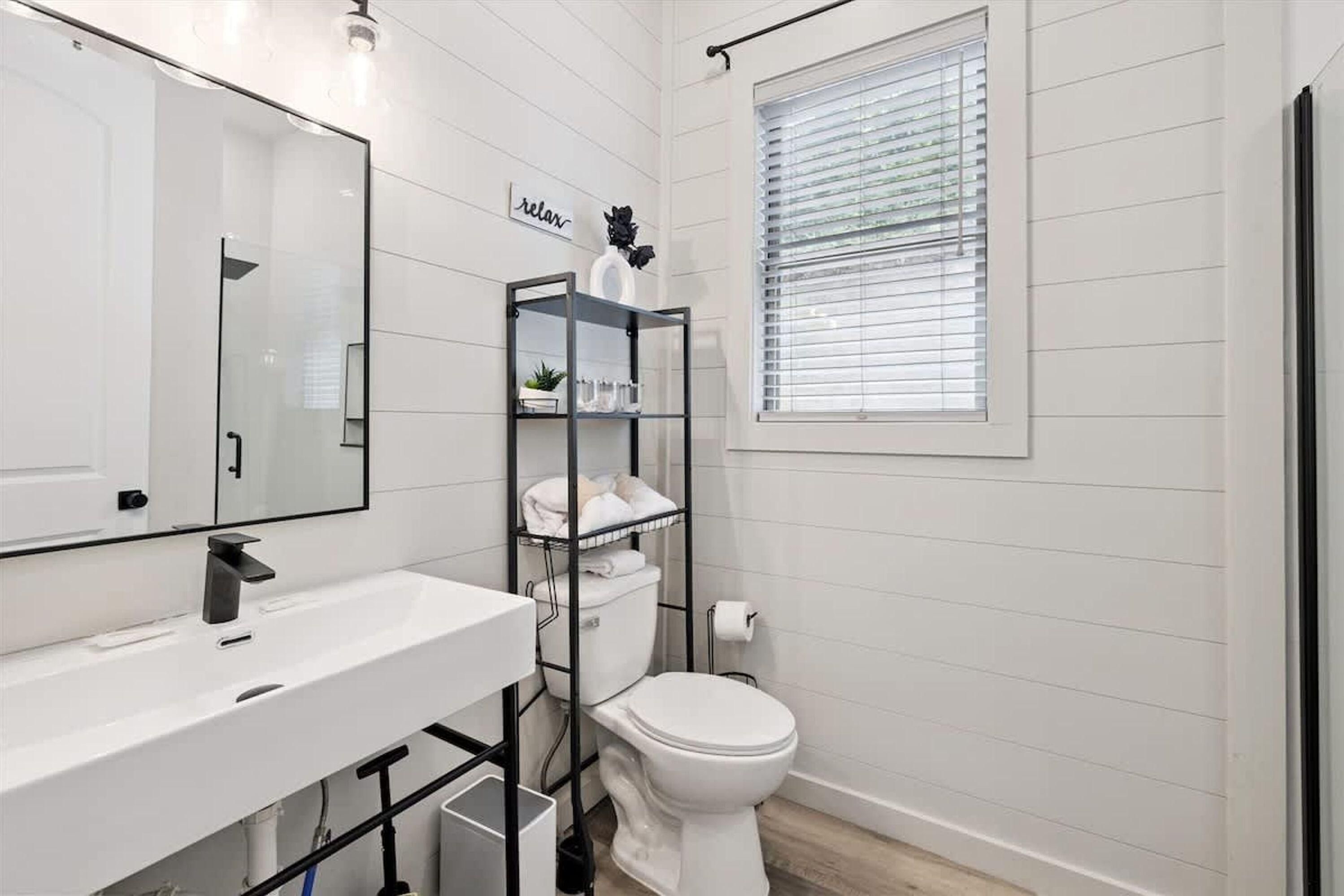Un baño moderno con un lavamanos blanco, un inodoro, una estantería de metal negro y una ventana con persianas.