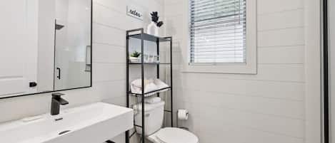 Un baño moderno con un lavamanos blanco, un inodoro, una estantería de metal negro y una ventana con persianas.