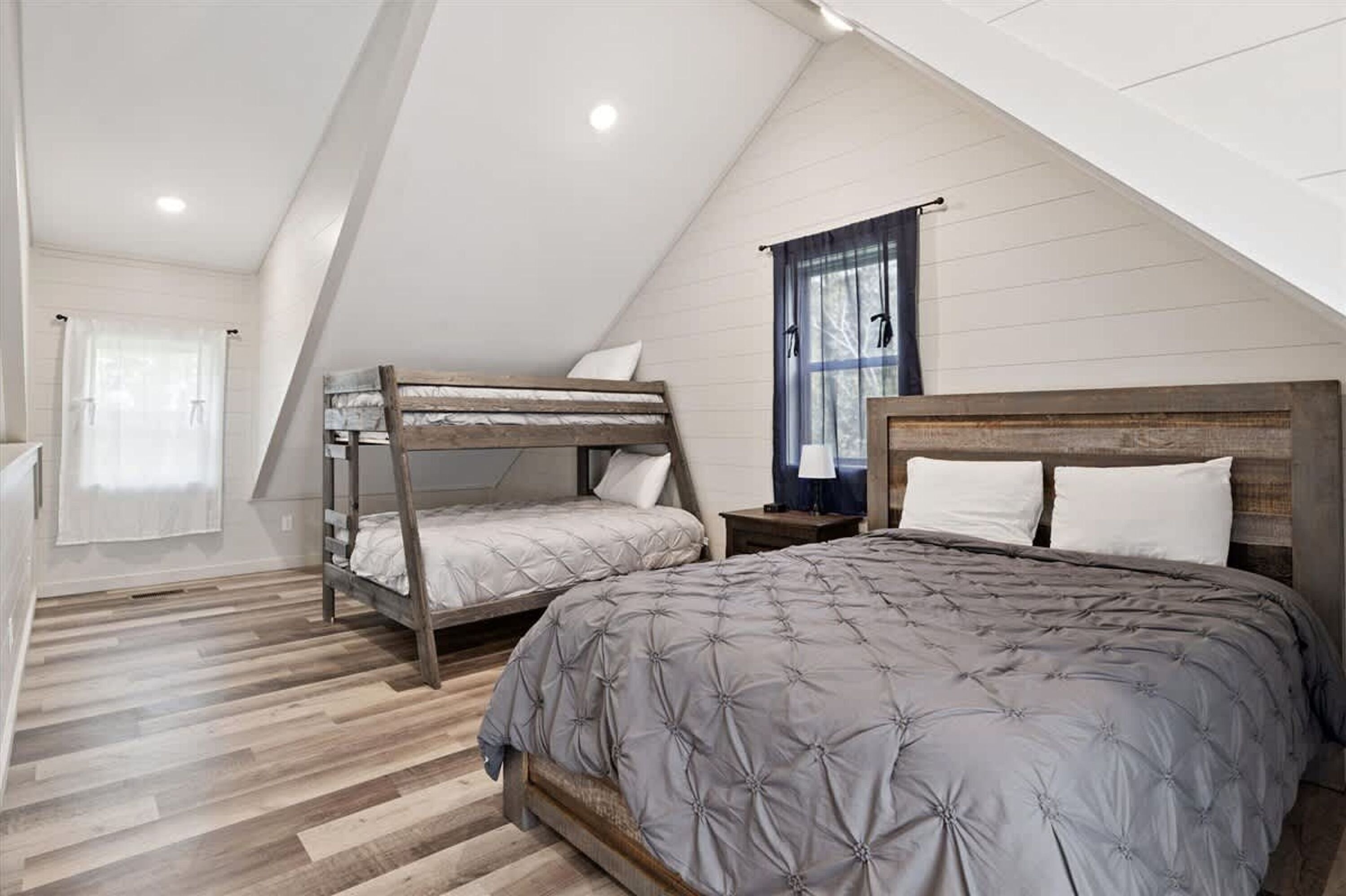 Un dormitorio con una cama litera, una cama individual, suelo de madera, una ventana con cortinas y una mesita de noche.