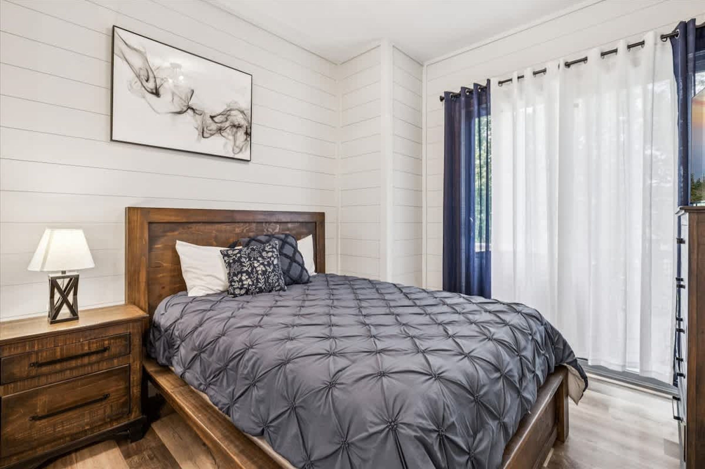 A bedroom with a bed, a nightstand, a lamp, a wall-mounted artwork, and a window with curtains.