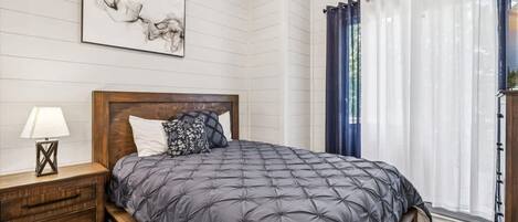 A bedroom with a bed, a nightstand, a lamp, a wall-mounted artwork, and a window with curtains.