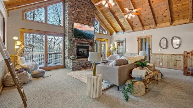 A spacious living room with a stone fireplace, large windows, and a high ceiling with exposed beams. - Round Top Way Cabin by Beyond Expectations (Sevierville)