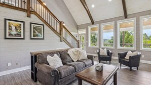 A spacious living room with a high ceiling, a wooden staircase, and large windows offering a view of the outdoors. - Mountain View Beauty by Beyond Expectations (Gatlinburg)