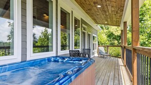 A wooden deck with a hot tub, outdoor seating, and large windows overlooking greenery. - Mountain View Beauty by Beyond Expectations (Gatlinburg)