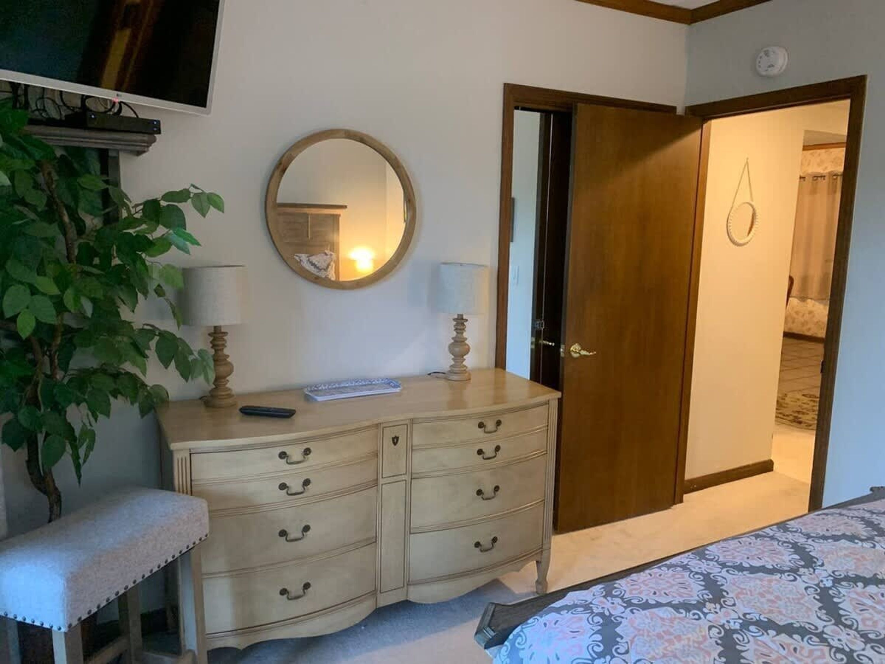 Une chambre à coucher comprenant un lit, une table de chevet, un miroir, une commode, une plante et une porte donnant sur une autre pièce.