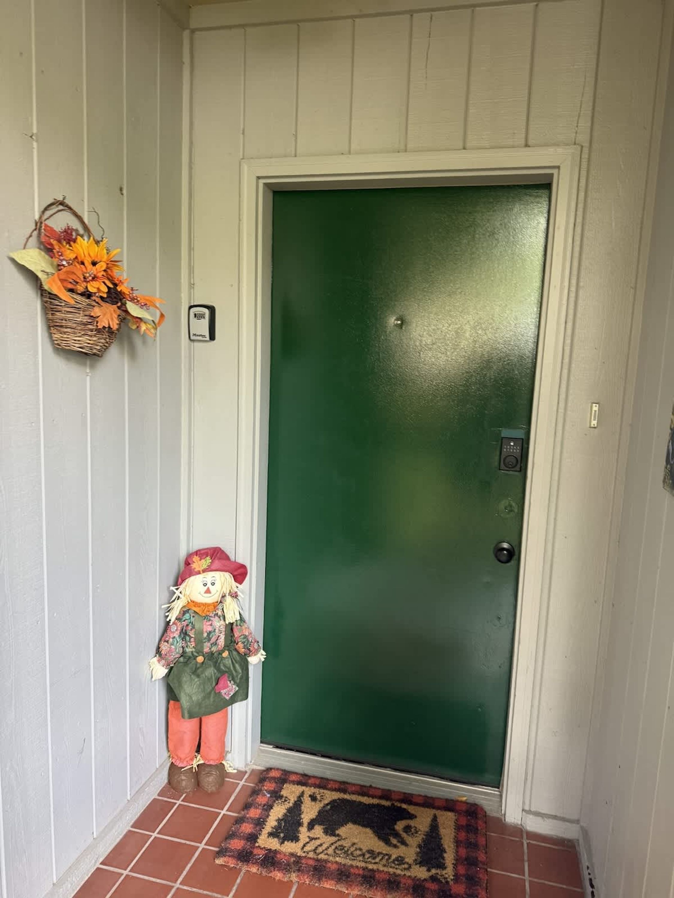 Una puerta verde con felpudo, una cesta decorativa con hojas otoñales y una figura de espantapájaros.