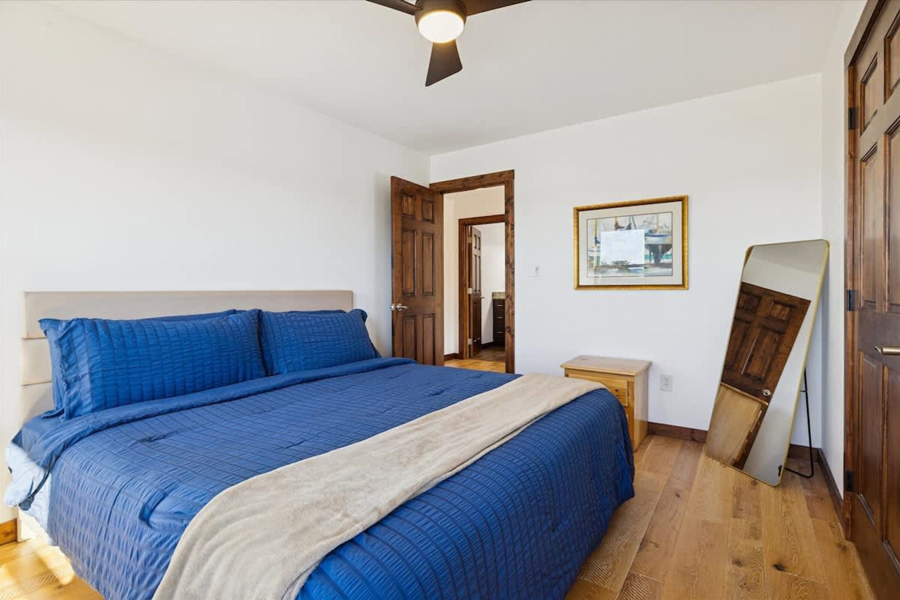 Quarto com uma cama grande, um ventilador de teto, uma porta de madeira e um quadro pendurado na parede.