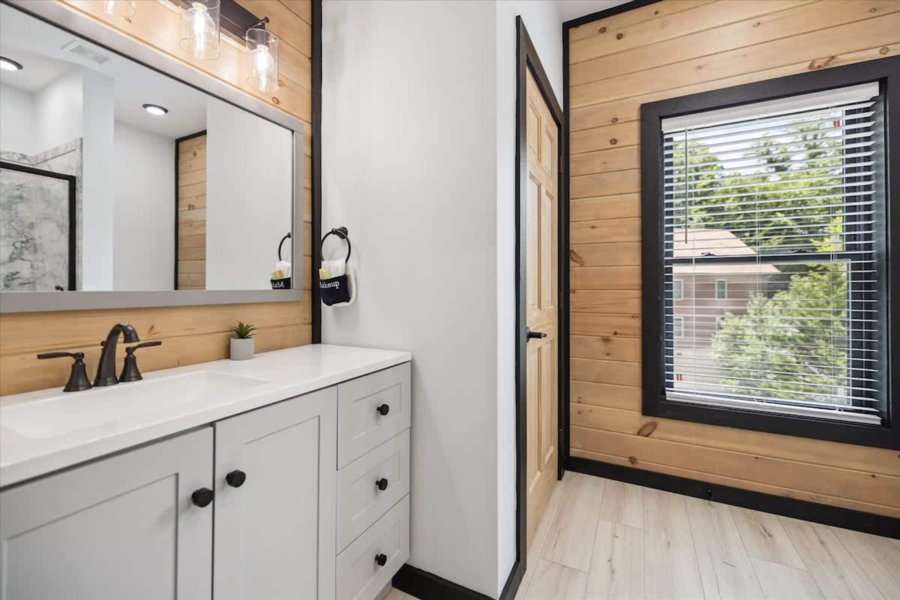 Une salle de bain moderne avec un meuble-lavabo blanc, un grand miroir et une fenêtre équipée de stores.