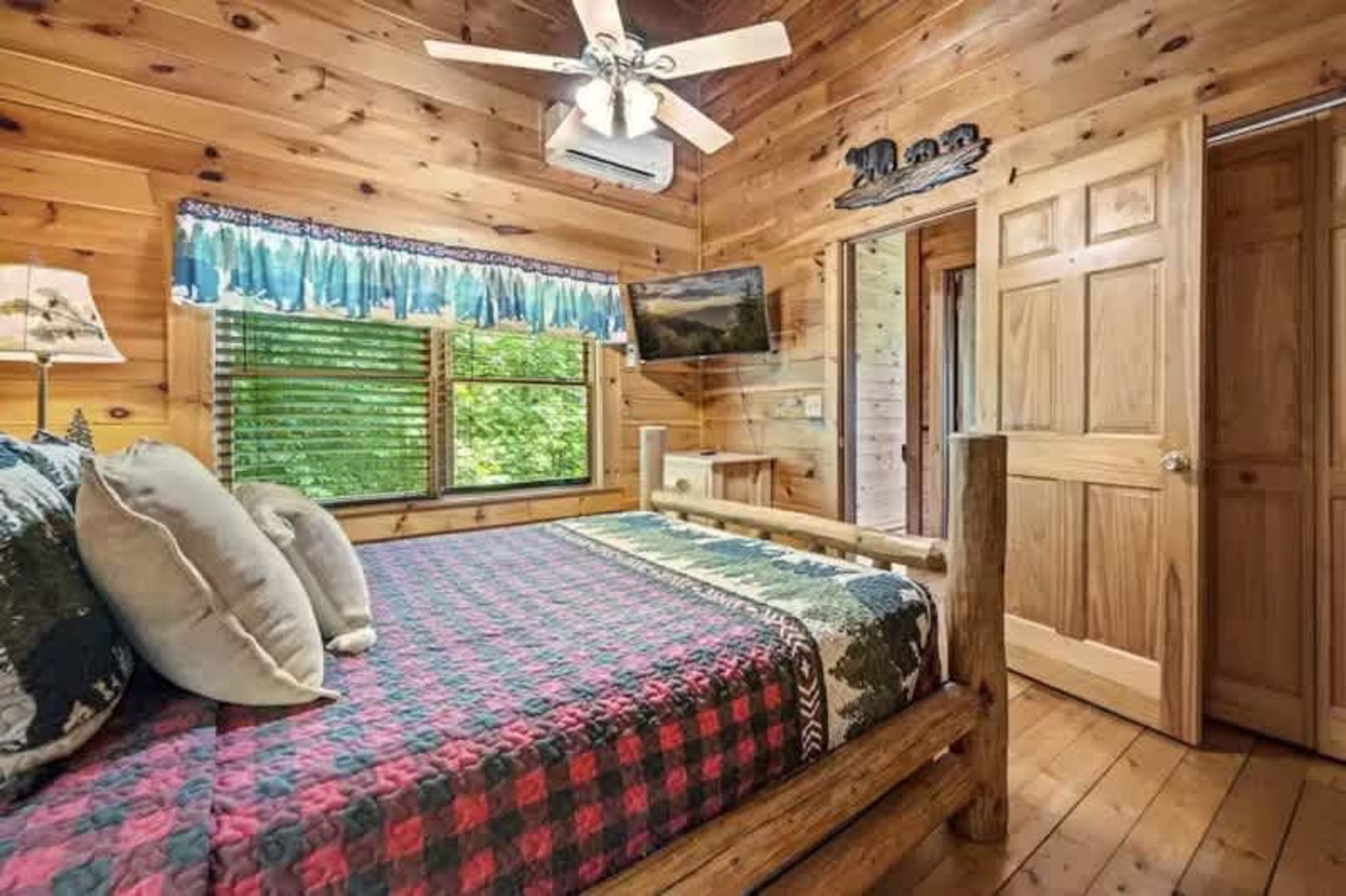 A wooden cabin bedroom with a bed, a ceiling fan, a TV, and a window with a view of trees.