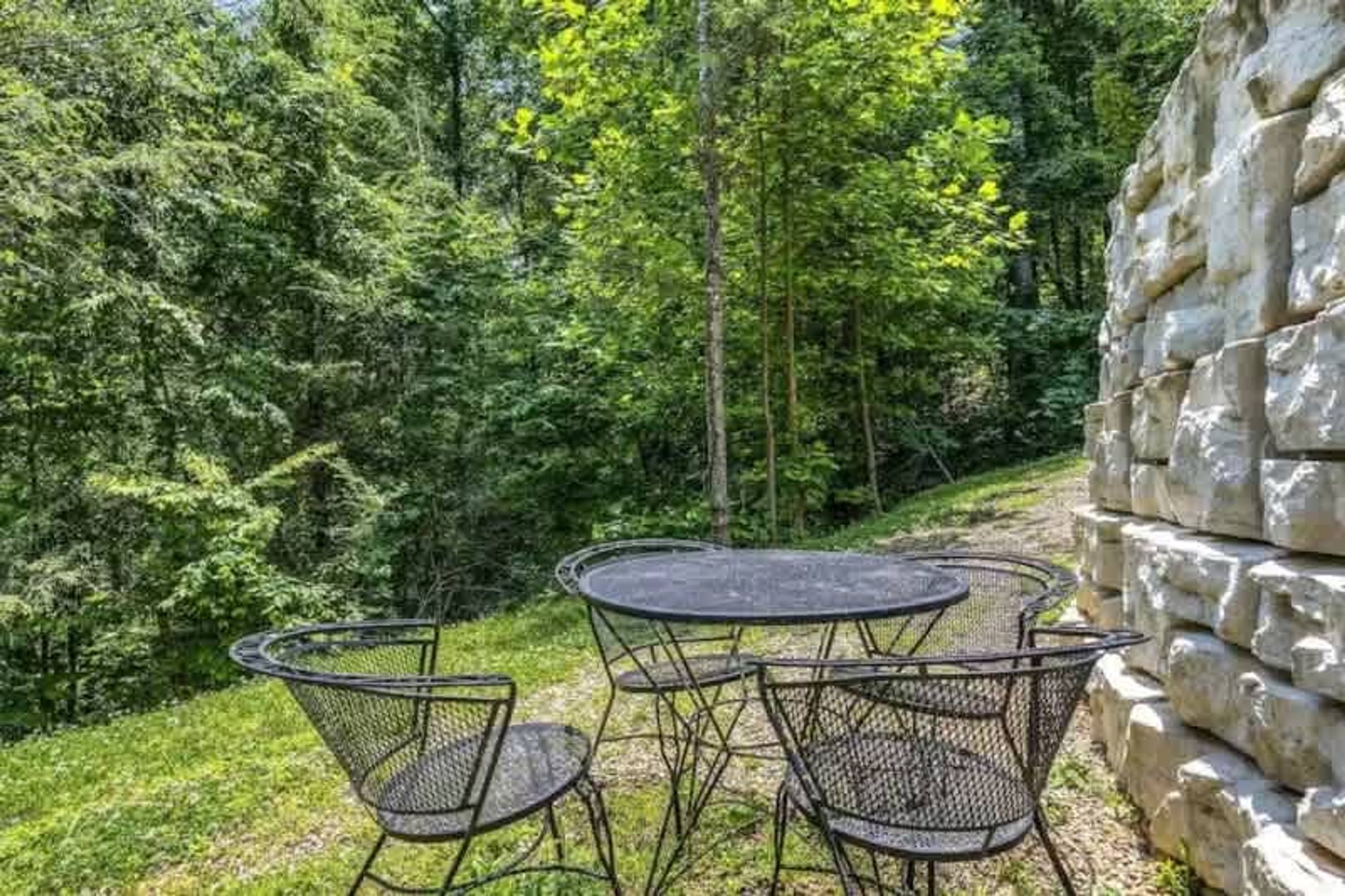 Juego de mesa y sillas de metal al aire libre, con abundante follaje verde y un muro de piedra de fondo.