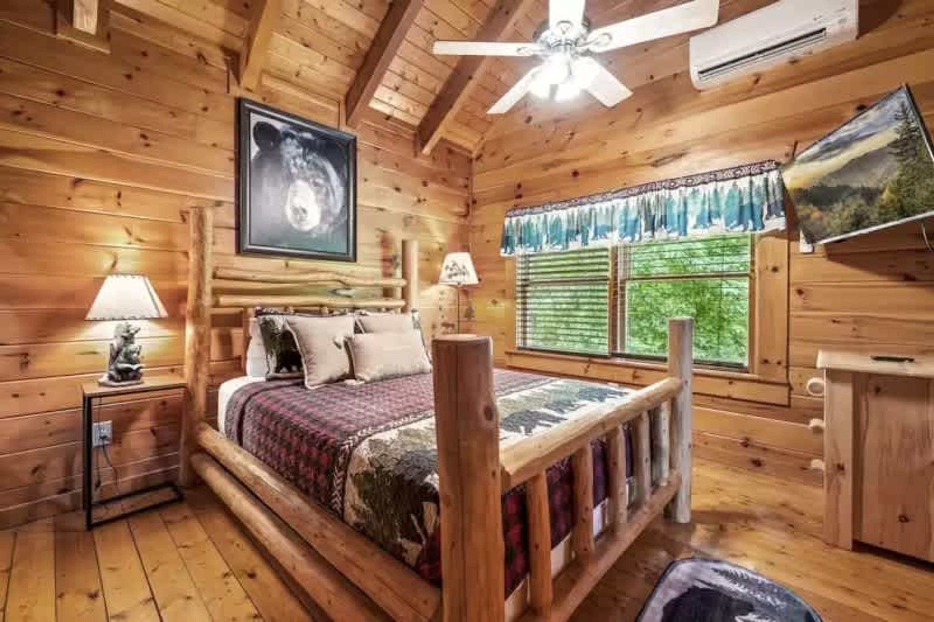 A wooden cabin bedroom with a bed, a ceiling fan, a window with curtains, and a wall-mounted TV.