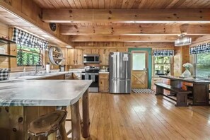 Interior de una cabaña de madera con cocina, comedor y un gran ventanal con vistas a árboles.