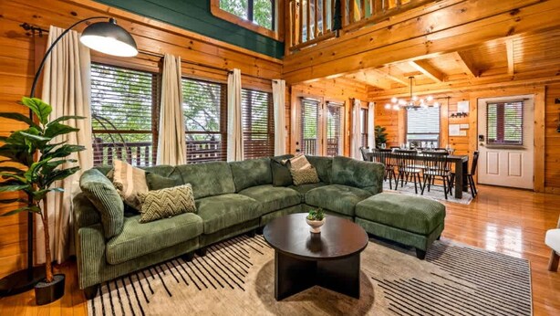 A cozy cabin interior with a green sectional sofa, a round coffee table, and a dining area with a table and chairs. - Smoky Haven Retreat by Beyond Expectations (Sevierville)