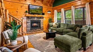 A cozy log cabin interior with a stone fireplace, green upholstered furniture, and a television mounted on the wall. - Smoky Haven Retreat by Beyond Expectations (Sevierville)