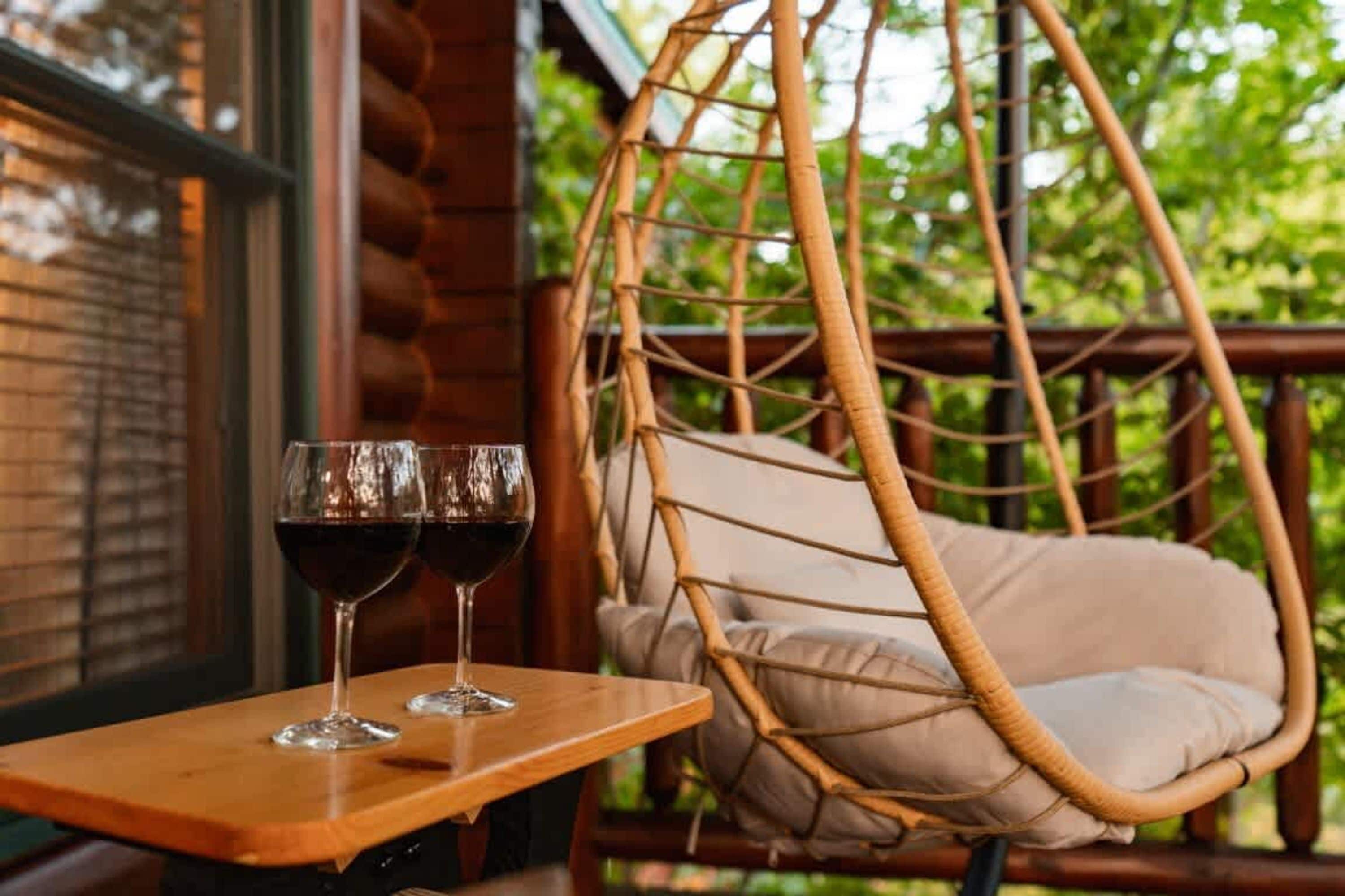 A wicker chair with a cushion, a wooden table with two wine glasses, and a window with blinds.
