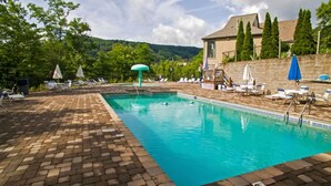 Una piscina rectangular con borde de ladrillo, rodeada de sillas de descanso y sombrillas, con árboles y un edificio de fondo.