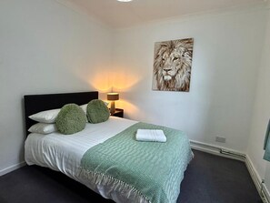 Una cama bien hecha con una colcha verde y blanca estampada, dos almohadas verdes y una toalla blanca. Se observa un cuadro colgado en la pared con un león.