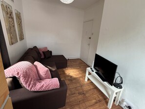 A living room with a sectional sofa, a flat-screen TV on a white stand, and a door in the background. - Clapham Road Apartment (Bedford)
