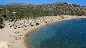 Ein Strand mit vielen Sonnenschirmen und Liegestühlen, ein sandiger Uferbereich und klares, blaues Wasser.