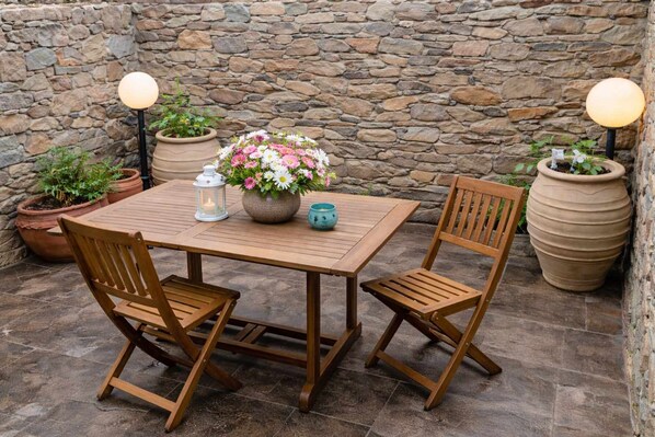 Une table en bois, deux chaises, un pot de fleurs et deux lampes de style lanterne, le tout placé devant un mur de pierre.