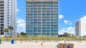 A multi-story hotel with balconies, located near a beach with sand and vegetation.