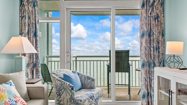 A room with a sofa, a chair, a table, a lamp, and a view of the sea through a sliding door.