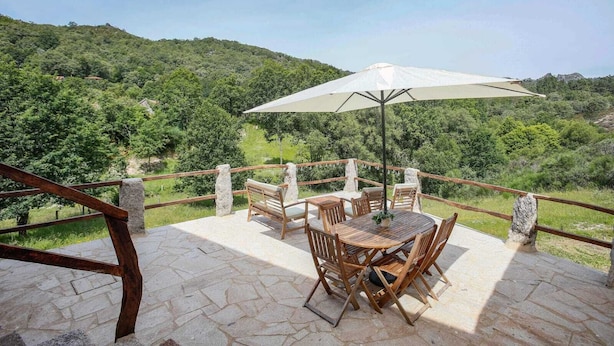 Una terraza de madera con una mesa redonda y sillas, un paraguas y una barandilla de piedra con vistas a un valle verde y exuberante.