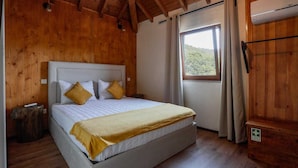 Una cama individual con cabecera blanca, dos almohadas y una manta amarilla. A la derecha, un banco de madera con un cartel verde, y una ventana con cortinas.