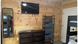 A compact room with a wooden wall, a flat-screen TV mounted above a dark wooden dresser, a metal shelf with folded clothes, and a bunk bed with a striped blanket. - Pole Barn Style Lodge located at Reelfoot Lake accommodating to large groups! (Hornbeak)