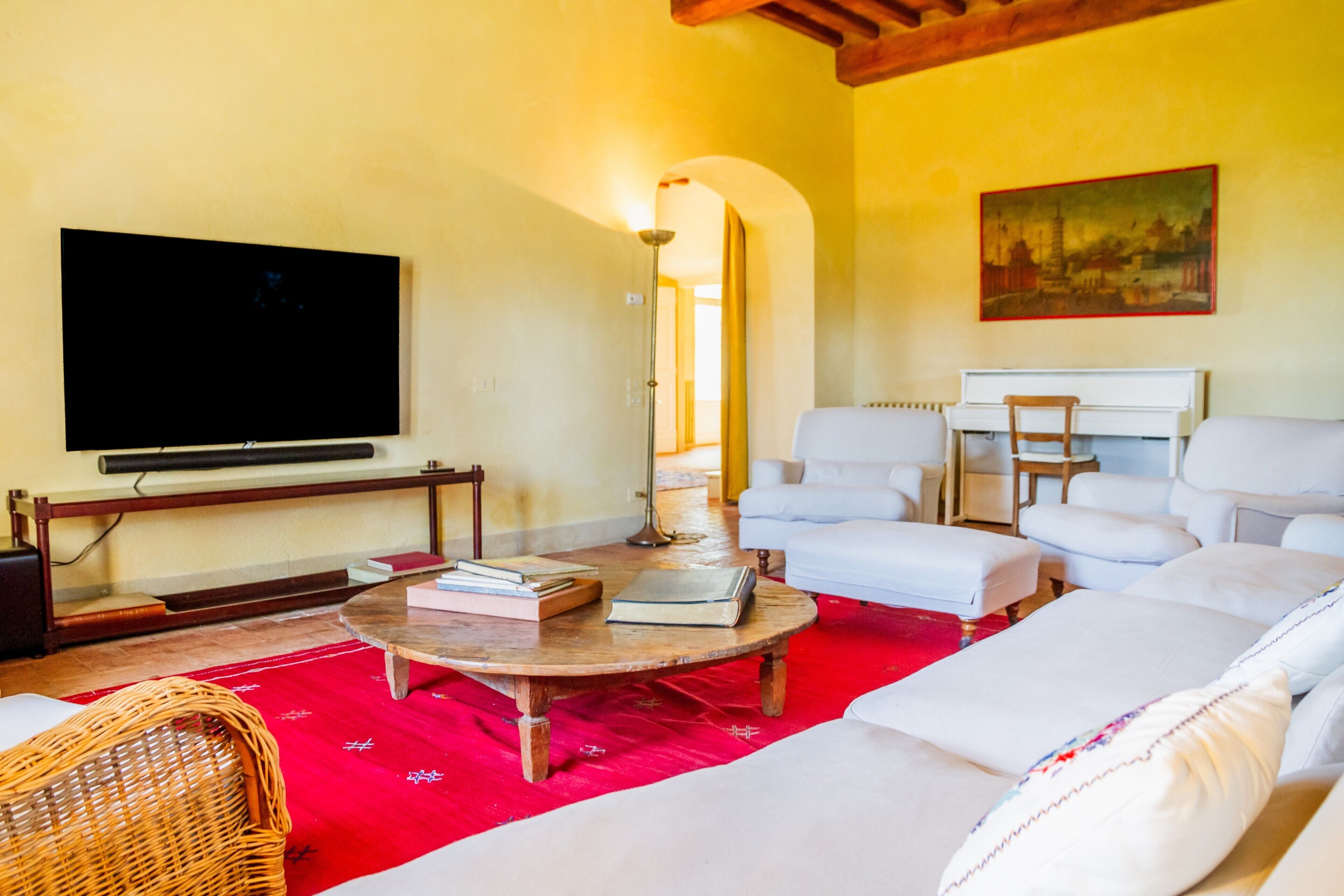 A living room with a flat-screen TV, a wooden coffee table, white sofas, a red rug, and a painting on the wall.