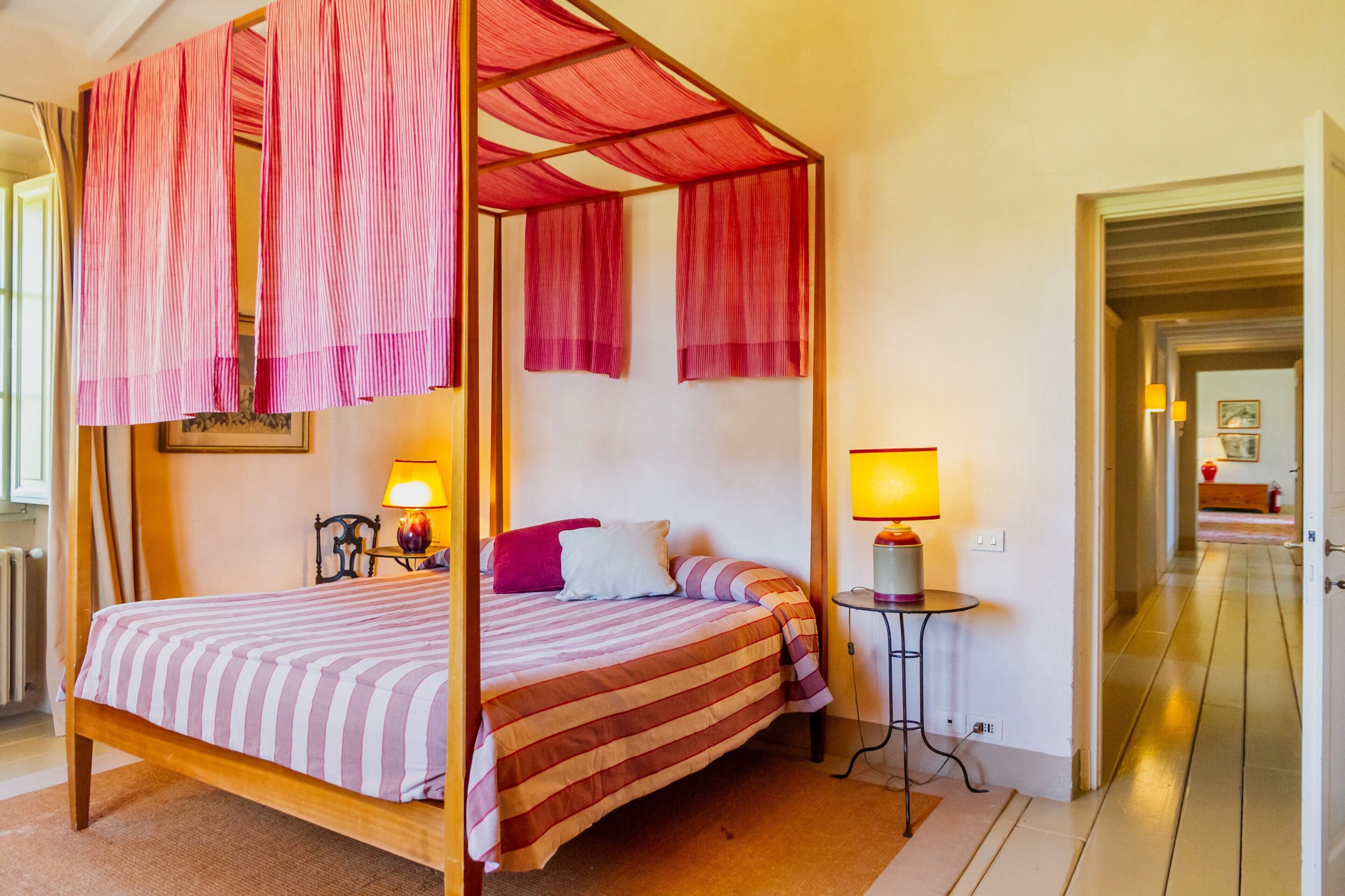 Een hemelbed met een gestreept laken, een nachtkastje met een lamp, en uitzicht op een gang.