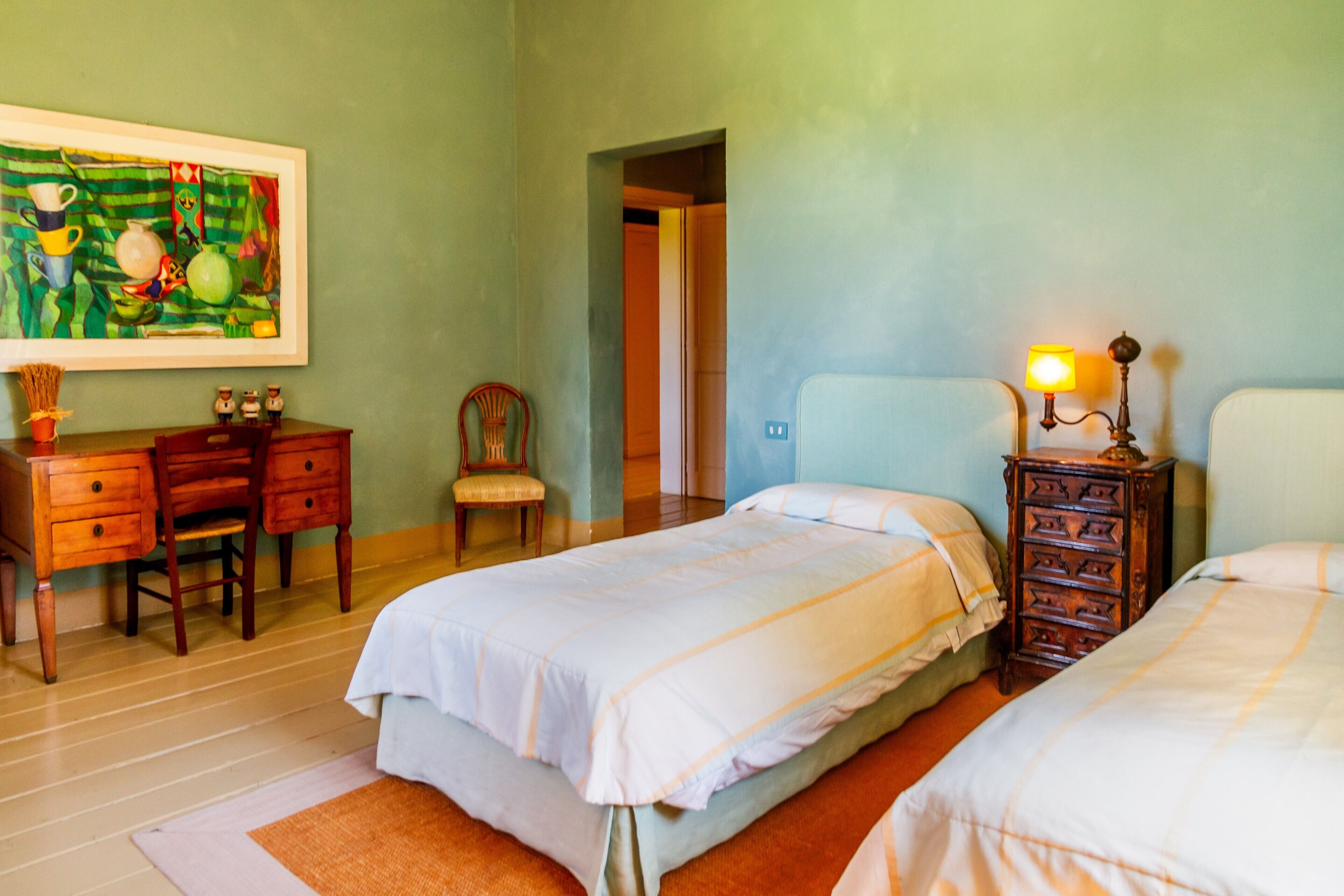 A bedroom with two beds, a desk with drawers, a chair, a lamp, and a painting on the wall.