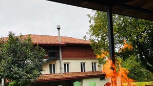 Un espacio al aire libre cubierto con una chimenea, sillas verdes y un edificio con techo de tejas rojas al fondo.