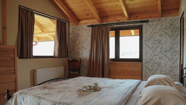 Un dormitorio con techo de madera, una cama con colcha estampada, dos ventanas con cortinas y un radiador.
