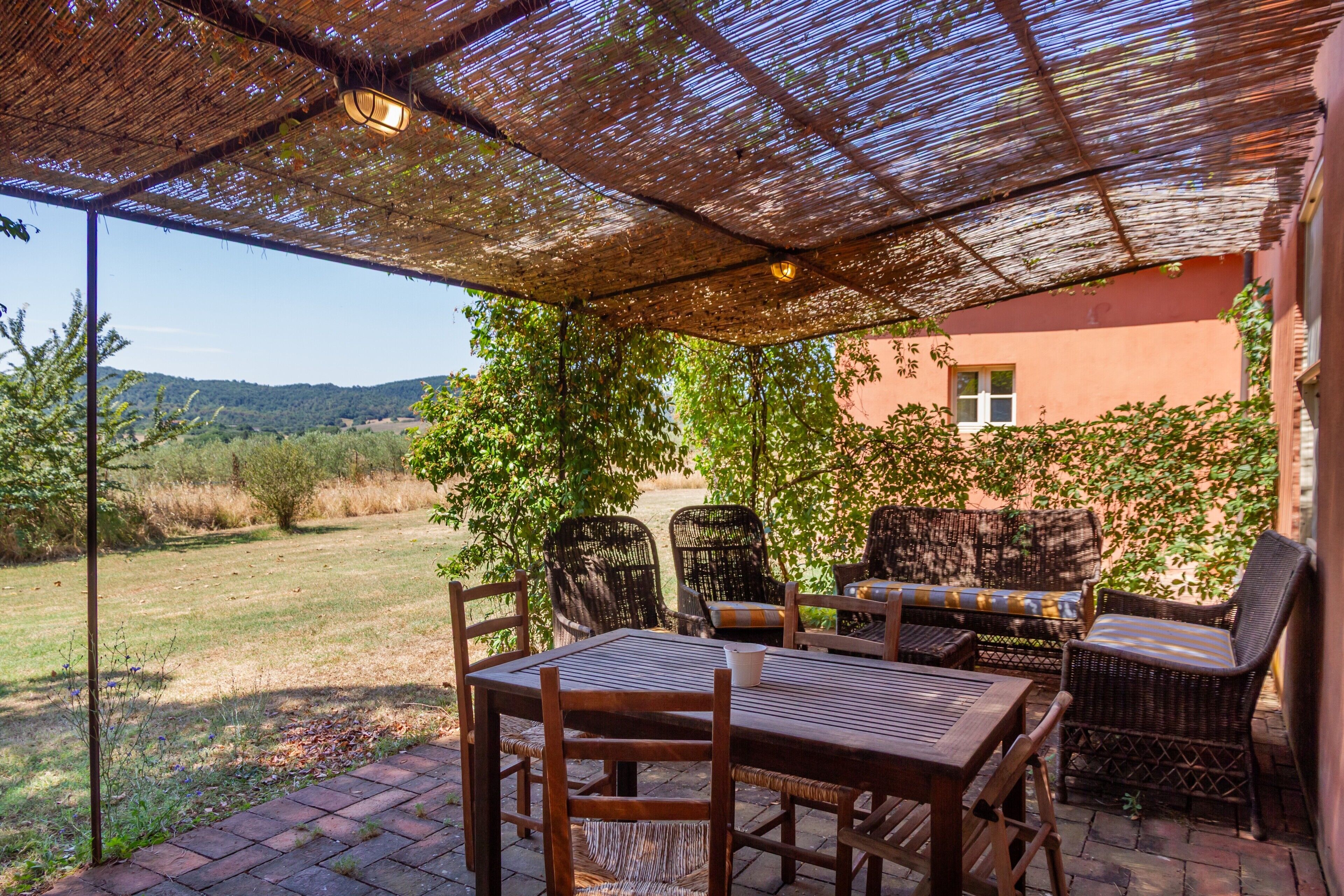 An outdoor patio with a table and chairs, a thatched roof, and a view of a green landscape.