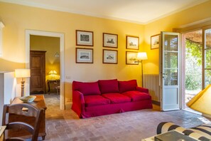 Ein Wohnzimmer mit einem roten Sofa, gerahmten Bildern an der Wand, einer Lampe und einer zur Außenseite führenden Tür.