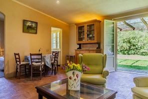 Ein Zimmer mit Esstisch, Stühlen, einer Couch, einem Glastisch mit Vase, einem Schrank und einer Glasschiebetür, die zu einem Garten führt.
