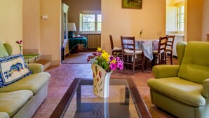 Una sala de estar con una mesa de centro de vidrio, un sofá, un sillón verde, una mesa de comedor con sillas y un cuadro en la pared.