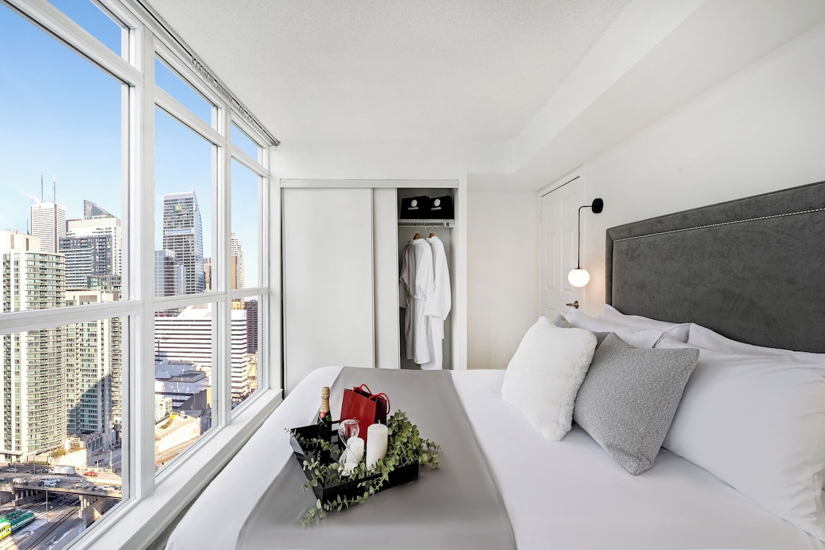 Ein Hotelzimmer mit großem Fenster, einem Bett mit weißer Bettwäsche, einem grauen Kopfteil und einem Nachttisch mit einer Blumenkomposition und einer Champagnerflasche.