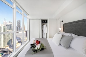 Ein Hotelzimmer mit großem Fenster, einem Bett mit weißer Bettwäsche, einem grauen Kopfteil und einem Nachttisch mit einer Blumenkomposition und einer Champagnerflasche.