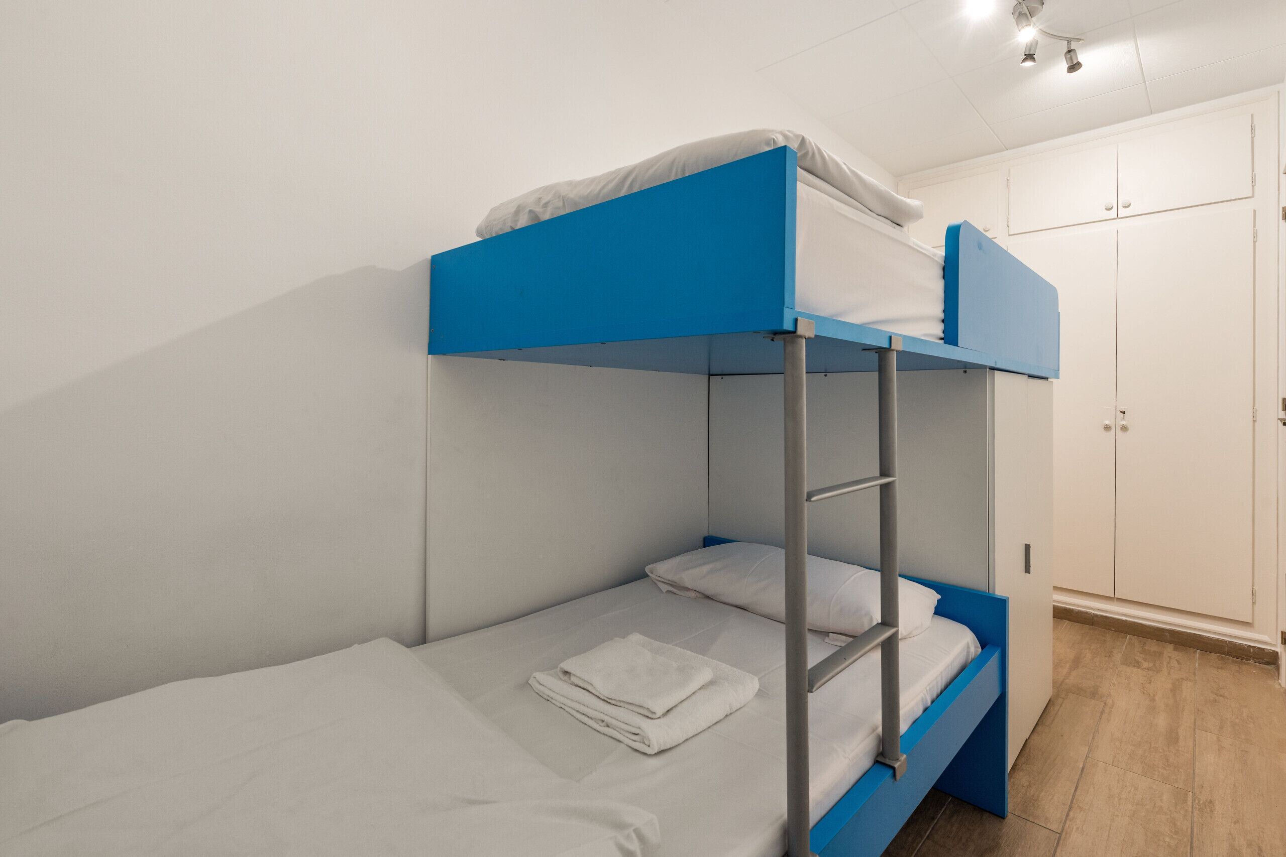 Una cama litera con estructura azul y ropa de cama blanca, un armario blanco y suelo de madera.