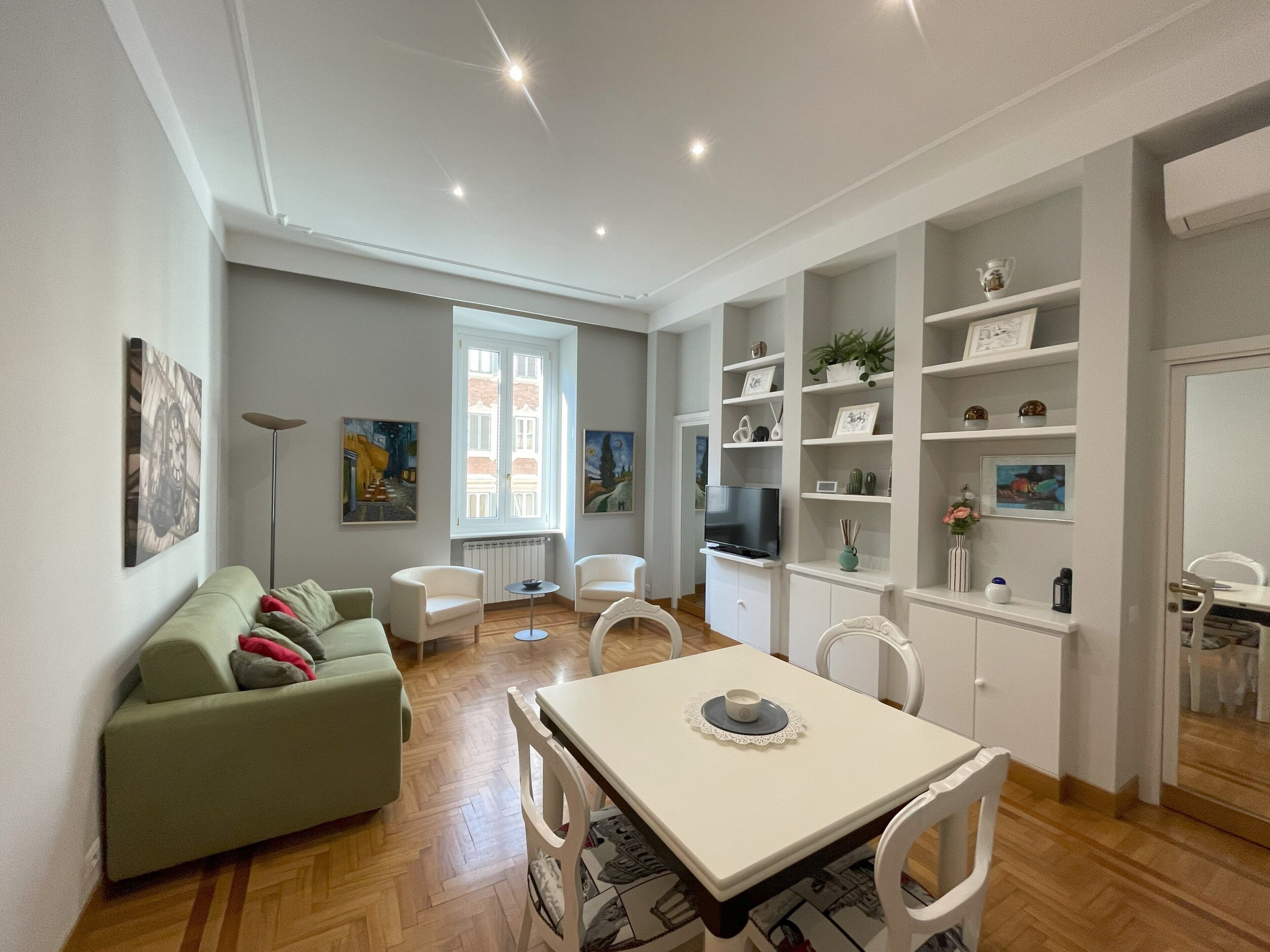 A modern living space with a dining area, sofa, TV, and shelves with various items.