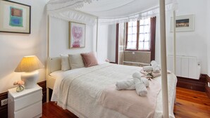 Un dormitorio con cama con dosel, una mesita de noche con lámpara, un cuadro enmarcado en la pared y una ventana con cortinas.