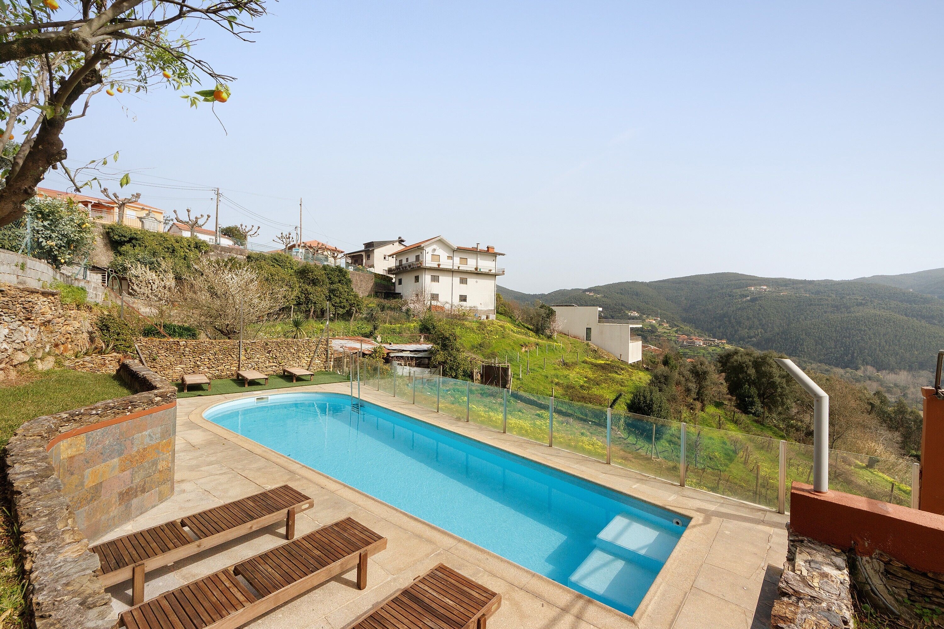Una piscina amb gandules, una paret de pedra i vista cap a cases i turons.