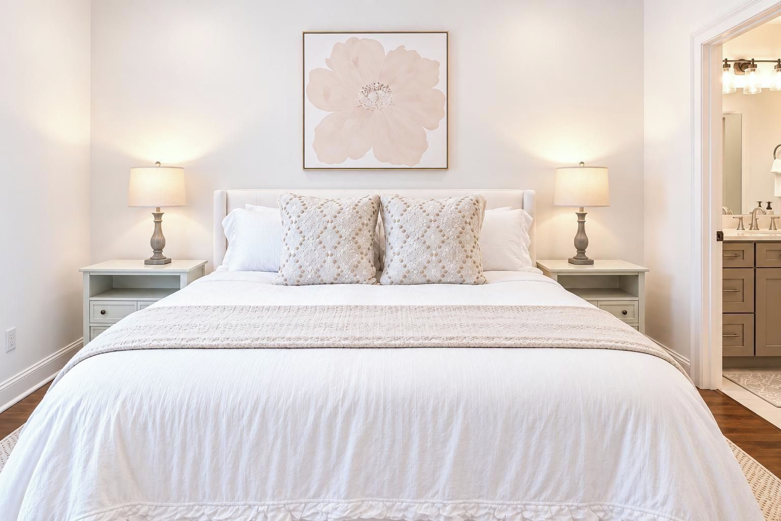 Een slaapkamer met een groot bed, twee nachtkastjes, een wandlampje en een bloemig kunstwerk boven het bed. Een badkamer is zichtbaar door een geopende deur.